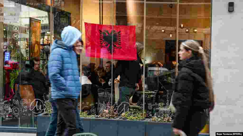 Disa persona kalojnë pranë një lokali në Prishtinë që ka vendosur në dritare një flamur kombëtar shqiptar. 